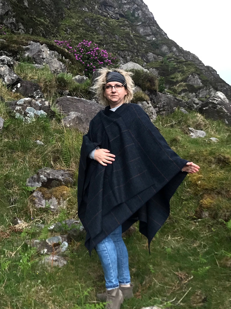 May include: A woman wearing a dark blue and black plaid poncho with a brown headband and jeans, standing in a grassy area with rocks and a mountain in the background.