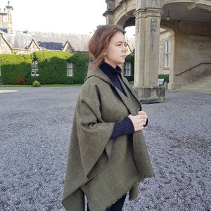 May include: A woman wearing a green hooded wool cape with a fringed edge. The cape is draped over her shoulders and she is standing in front of a stone building.