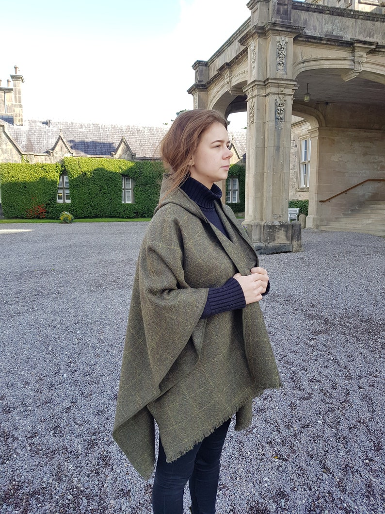 May include: A woman wearing a green hooded wool cape with a fringed edge. The cape is draped over her shoulders and she is standing in front of a stone building.