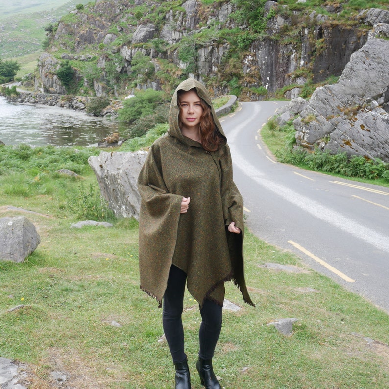 May include: A green hooded poncho with a fringed edge. The poncho is made of a tweed fabric and is worn by a person standing on a paved road.