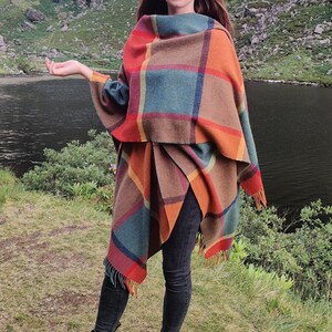 May include: A woman wearing a colorful plaid shawl stands in a grassy field with a lake and mountains in the background. She is wearing black jeans and black boots.