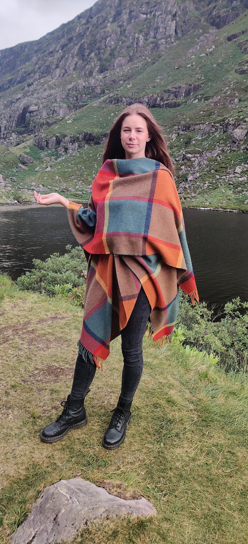 May include: A woman wearing a colorful plaid shawl stands in a grassy field with a lake and mountains in the background. She is wearing black jeans and black boots.