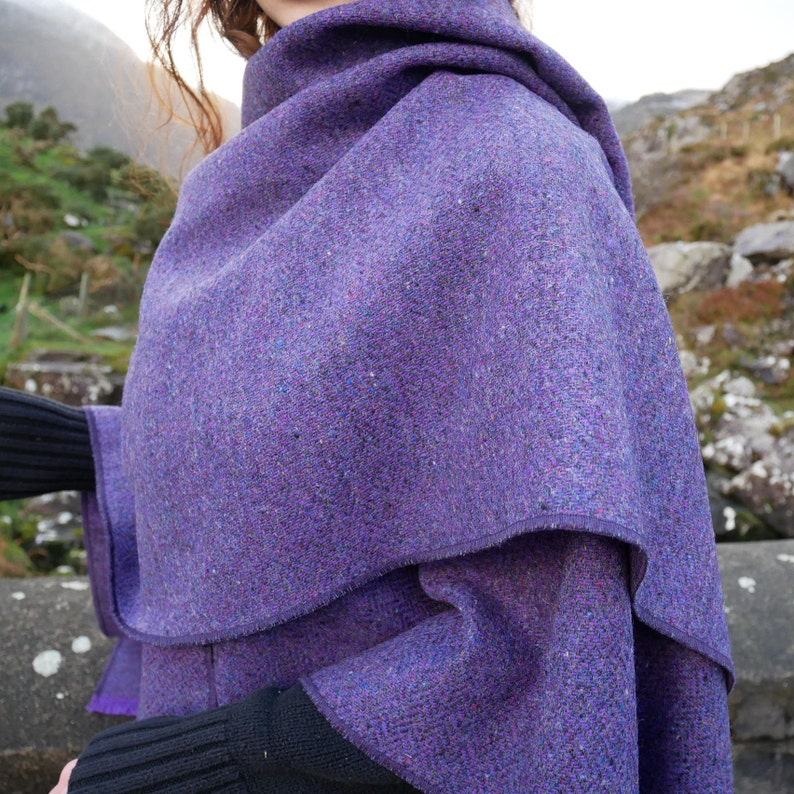 May include: A purple tweed shawl with a layered design. The shawl is a vibrant shade of purple with flecks of other colors. The fabric appears to be thick and warm, suitable for cooler weather. The shawl is draped over the shoulders.