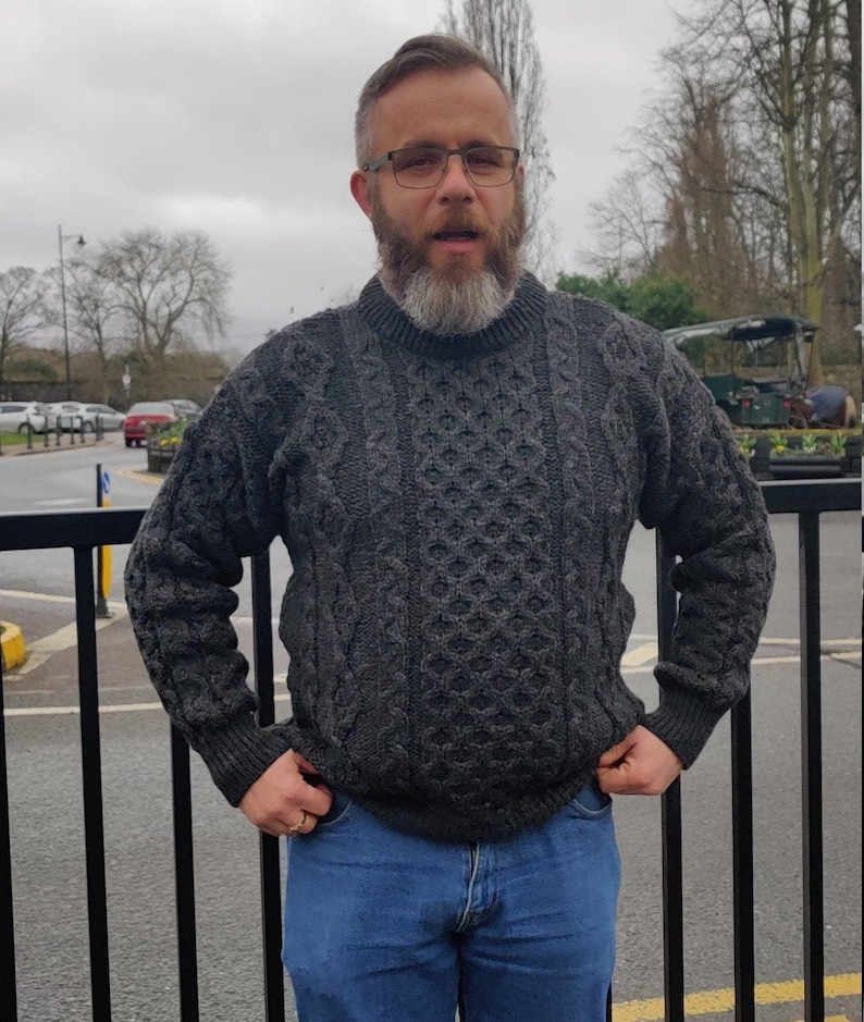 May include: A man wearing a gray cable knit sweater and blue jeans. He has a beard and is wearing glasses.