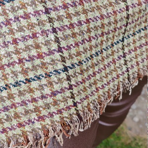 May include: A brown, green, and white plaid fabric with a frayed edge. The fabric is woven with a small checkered pattern.