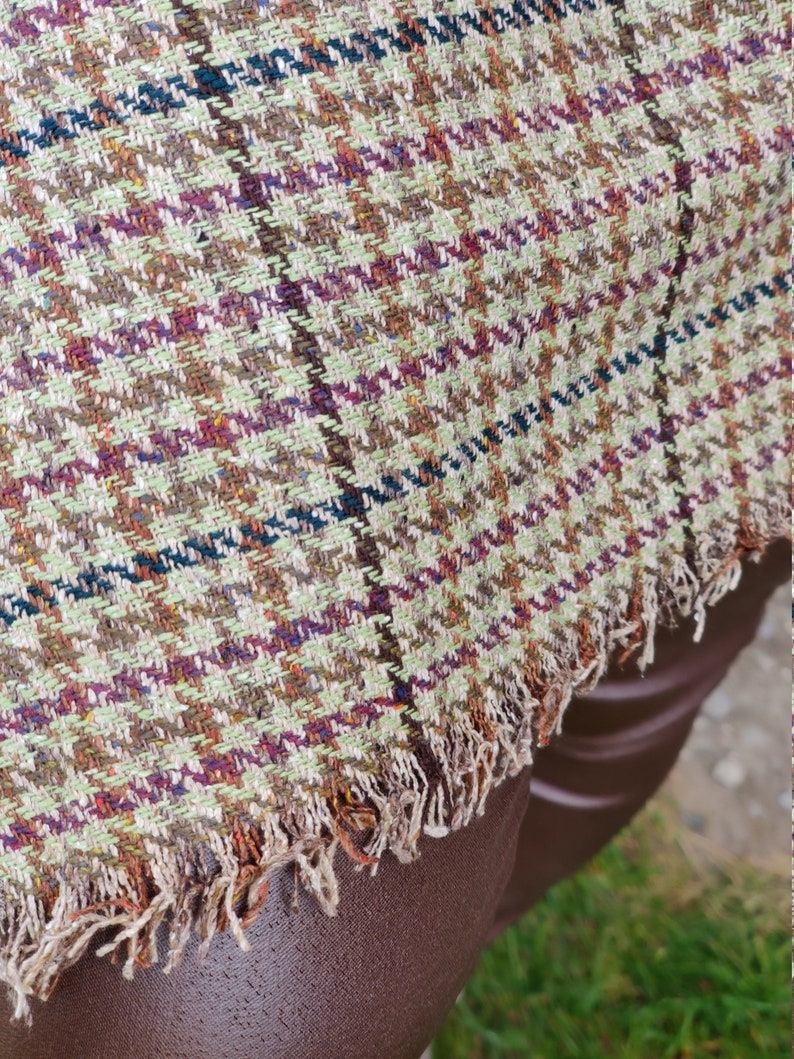 May include: A brown, green, and white plaid fabric with a frayed edge. The fabric is woven with a small checkered pattern.