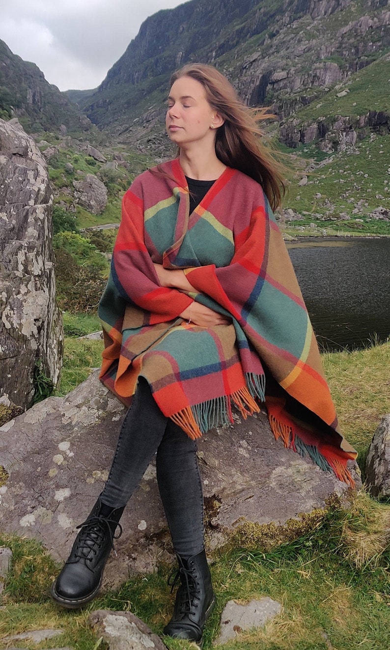 May include: A woman wearing black pants and black boots sits on a rock with a colorful plaid shawl wrapped around her shoulders. The shawl has a fringe border and is made of a soft, woven fabric.