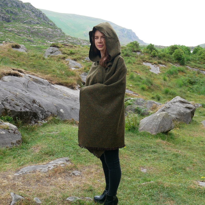 May include: A woman wearing a green hooded wool shawl with a fringe trim. She is standing in a grassy field with rocks in the background.