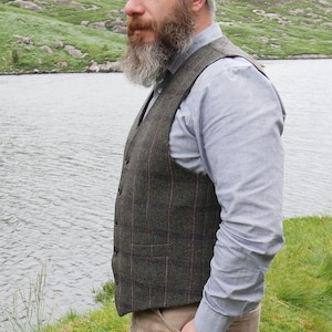 Peut inclure: Un homme portant un gilet &agrave; carreaux vert et rouge sur une chemise boutonn&eacute;e bleu clair. Il a une longue barbe grise et se tient devant un lac.