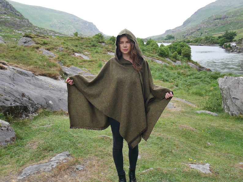 May include: A green hooded poncho with a fringed edge. The poncho is made of a soft, textured fabric and is worn by a person standing in a grassy field.