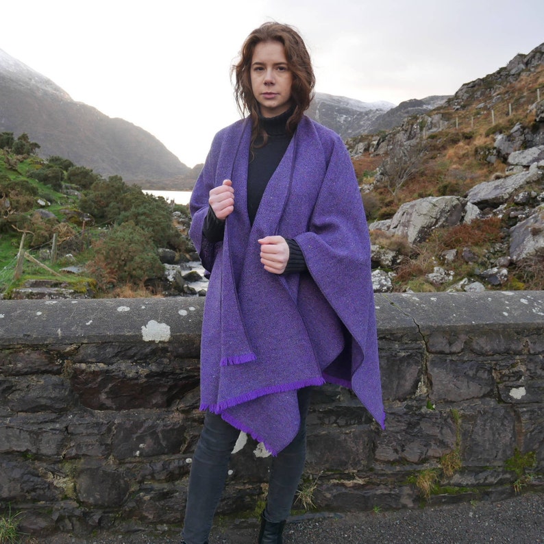 May include: A woman wearing a vibrant purple cape with a fringed hem. The cape is a solid color and appears to be made of a woven material. The background features a stone wall, mountains, and a body of water.