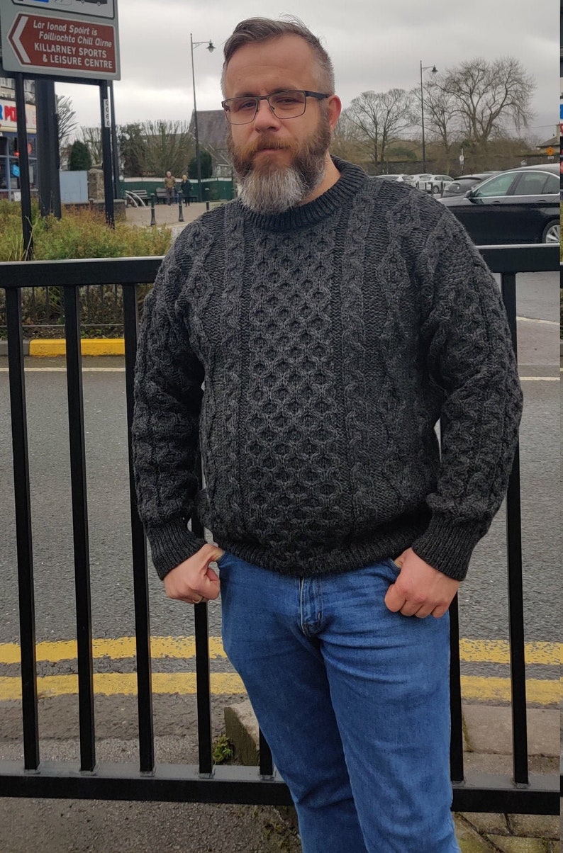 May include: A man wearing a gray cable knit sweater and blue jeans. He is standing in front of a black metal fence.