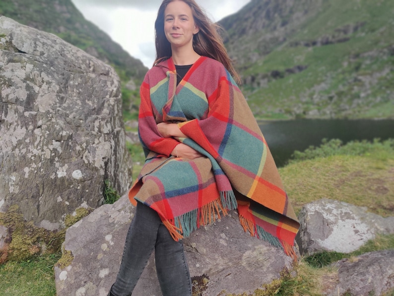 May include: A woman wearing a colorful plaid poncho with fringe, sitting on a large rock in a natural setting.