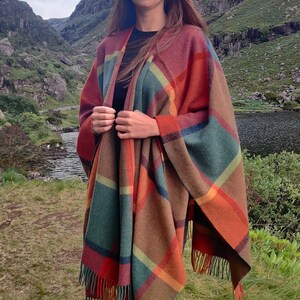 May include: A woman wearing a colorful plaid poncho with fringe, black jeans, and black boots. She is standing in a grassy field with a lake and mountains in the background.