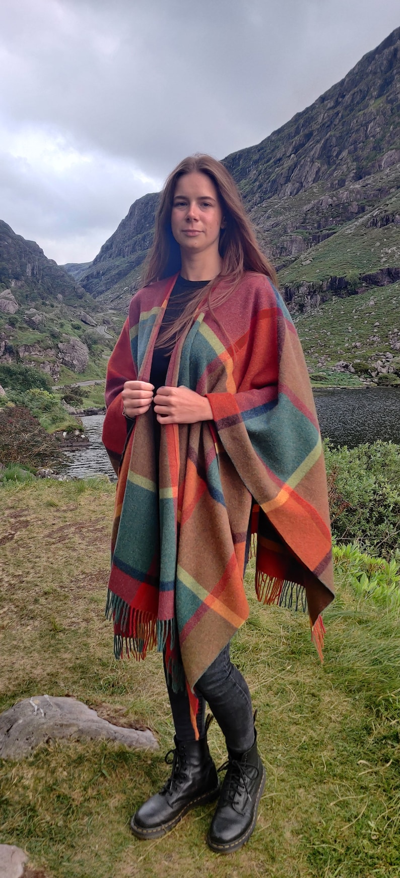 May include: A woman wearing a colorful plaid poncho with fringe, black jeans, and black boots. She is standing in a grassy field with a lake and mountains in the background.