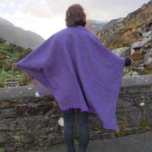 May include: A person wearing a vibrant purple poncho with a fringed hem, standing outdoors. The poncho is made of a thick, textured fabric. The background features a stone wall, mountains, and a cloudy sky.