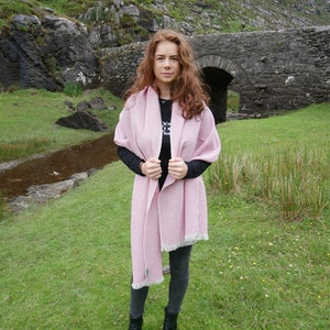 May include: A pink wool shawl with a fringe border. The shawl is draped around a person standing in front of a stone bridge.