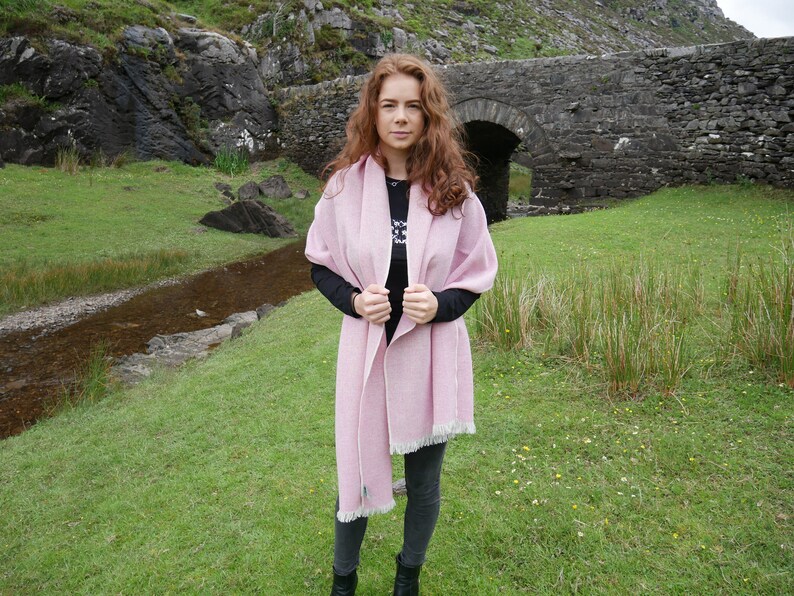 May include: A pink wool shawl with a fringe border. The shawl is draped around a person standing in front of a stone bridge.