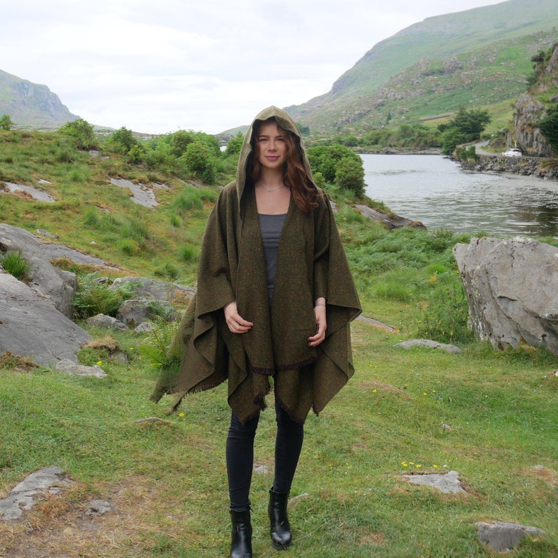 May include: A woman wearing a green hooded cloak with a fringed edge. She is standing in a grassy field with a river and mountains in the background.