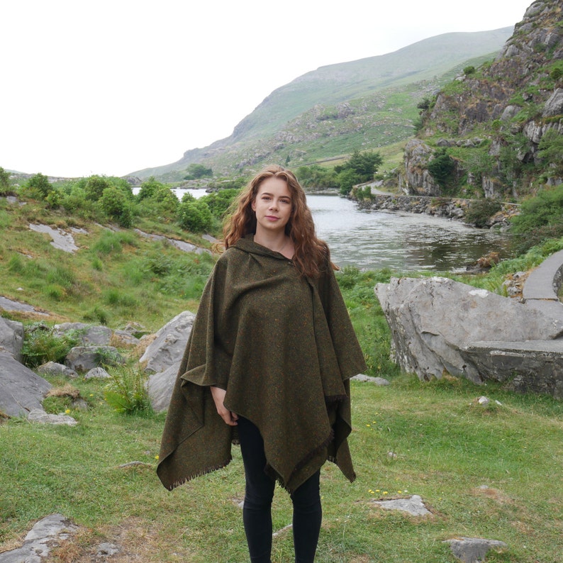 May include: A woman wearing a green hooded poncho standing in a grassy field with a river and mountains in the background.