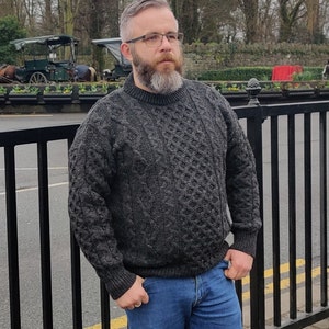 May include: A man wearing a dark gray cable knit sweater with a crew neck. He is wearing blue jeans and has a beard and glasses.