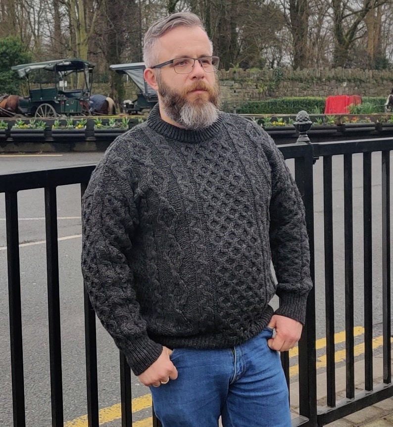 May include: A man wearing a dark gray cable knit sweater with a crew neck. He is wearing blue jeans and has a beard and glasses.
