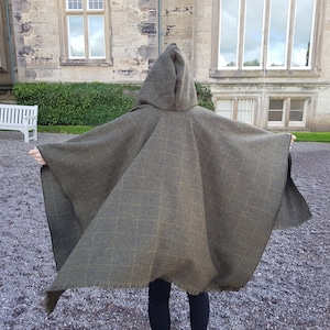 May include: A hooded green and brown plaid cape with a fringed edge. The cape is worn by a person standing in front of a stone building.