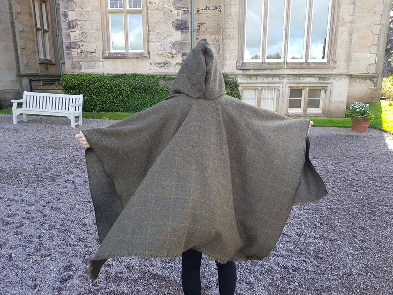May include: A hooded green and brown plaid cape with a fringed edge. The cape is worn by a person standing in front of a stone building.
