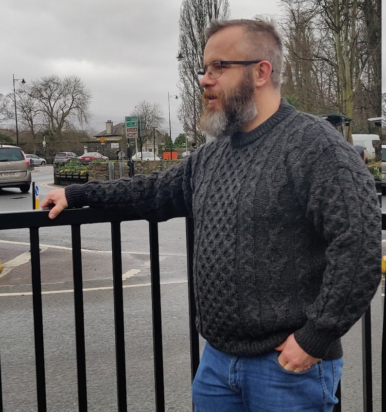 May include: A man wearing a dark gray cable knit sweater with a beard and glasses. He is standing in front of a black metal fence.