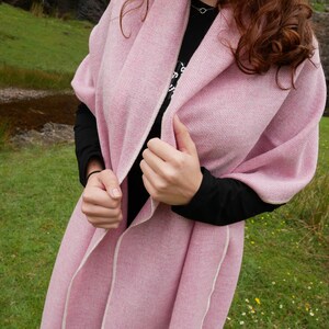 May include: A pink knitted shawl with a white trim. The shawl is draped over a person's shoulders and is being held in place by their hands.