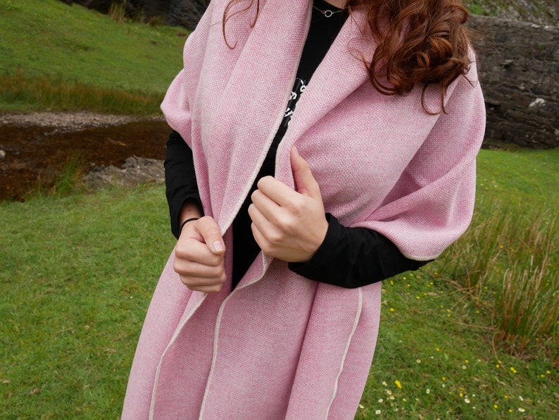 May include: A pink knitted shawl with a white trim. The shawl is draped over a person's shoulders and is being held in place by their hands.