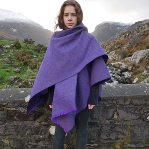 May include: A purple tweed cape with a hood and fringed edges. The cape is layered, with a darker purple lining visible at the edges. The person is wearing black clothing. The background features a stone wall and a mountainous landscape.