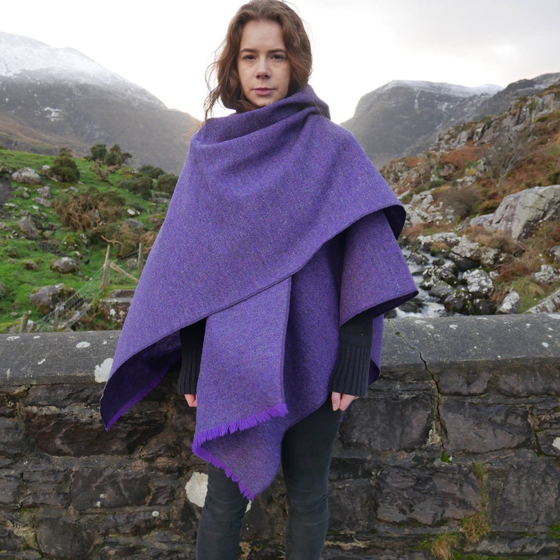 May include: A purple tweed cape with a hood and fringed edges. The cape is layered, with a darker purple lining visible at the edges. The person is wearing black clothing. The background features a stone wall and a mountainous landscape.