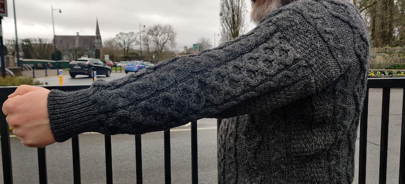 May include: A gray cable knit sweater with a textured pattern. The sweater is worn by a person with their arm extended.