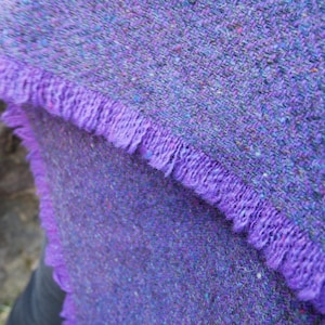 May include: Close-up of a purple tweed scarf with a fringed edge. The scarf has a textured appearance with flecks of color throughout. The fringe is a solid, vibrant purple.