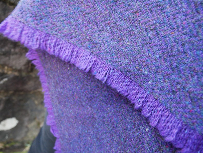 May include: Close-up of a purple tweed scarf with a fringed edge. The scarf has a textured appearance with flecks of color throughout. The fringe is a solid, vibrant purple.