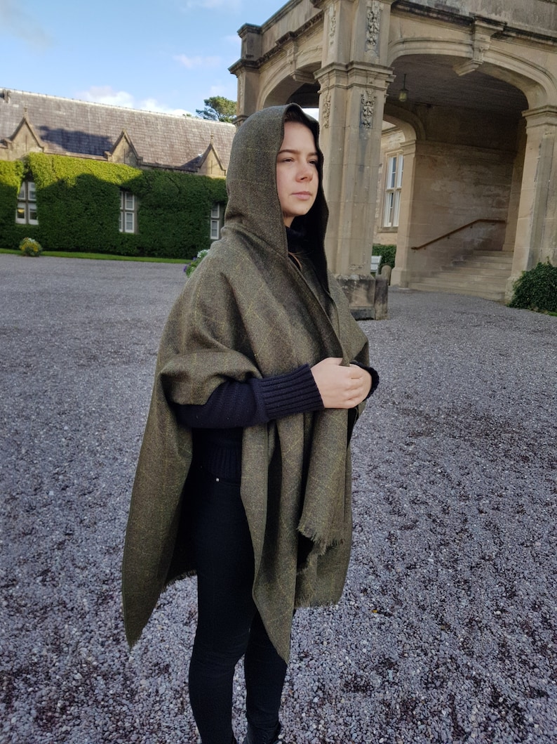 May include: A person wearing a green hooded shawl with a black sweater and black pants. The shawl has a plaid pattern and is draped over the person's shoulders. The person is standing in front of a stone building.