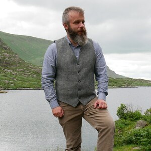 May include: A man wearing a gray plaid vest over a light blue button-down shirt. He is standing in front of a lake with a mountain in the background.