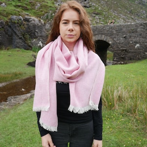 May include: A woman wearing a pink and white scarf with a fringed edge. The scarf is wrapped around her neck and tied in a loose knot. She is standing in front of a stone bridge and a green grassy field.