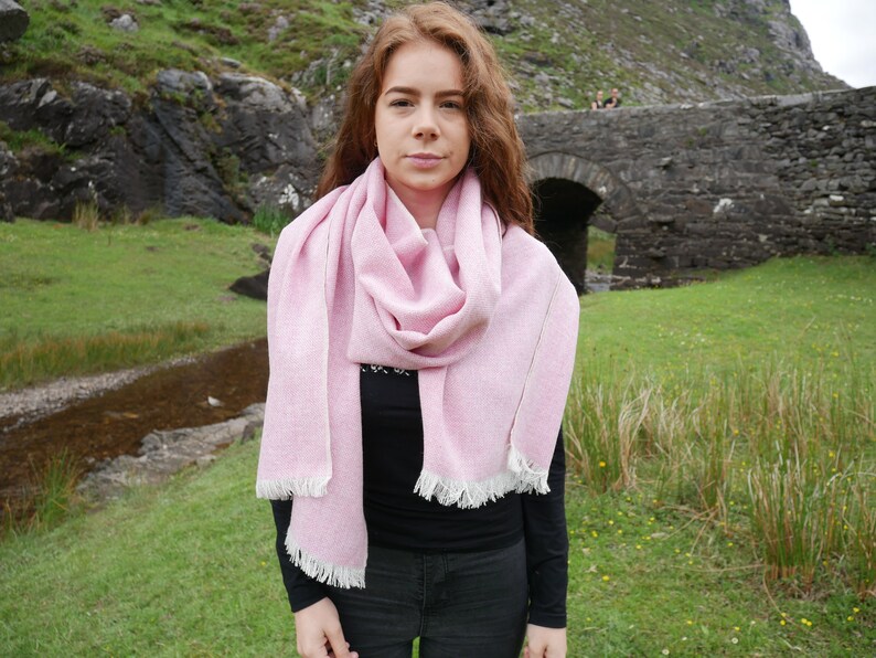 May include: A woman wearing a pink and white scarf with a fringed edge. The scarf is wrapped around her neck and tied in a loose knot. She is standing in front of a stone bridge and a green grassy field.