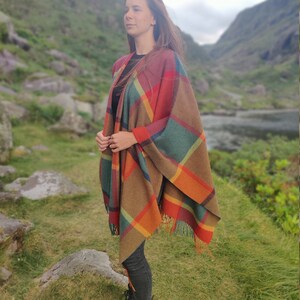 May include: A woman wearing a colorful plaid poncho with fringe. The poncho is brown, orange, red, green, and blue. She is standing in a grassy field with mountains in the background.