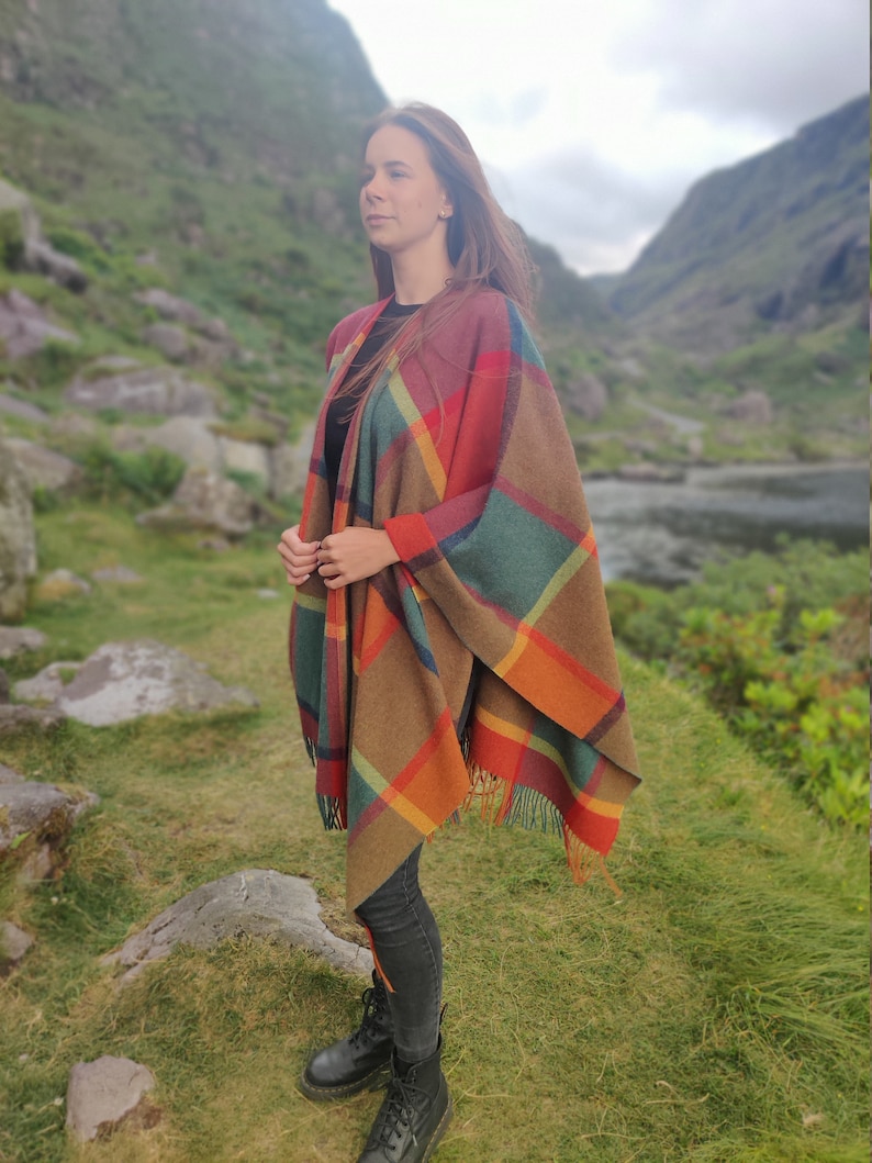 May include: A woman wearing a colorful plaid poncho with fringe. The poncho is brown, orange, red, green, and blue. She is standing in a grassy field with mountains in the background.
