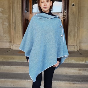 Könnte beinhalten: Ein hellblauer Strickponcho mit weißem Saum und drei silbernen Knöpfen. Der Poncho ist über die Schultern einer Person gelegt und wird mit einer schwarzen Hose getragen.