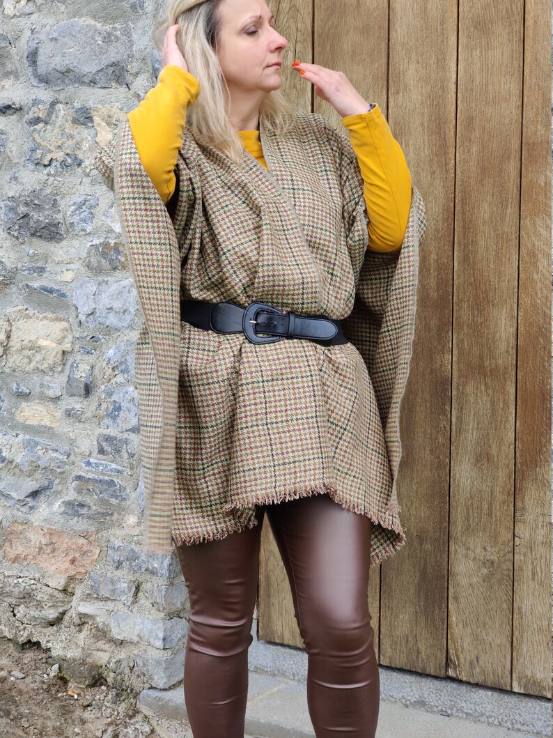 May include: A brown and tan plaid poncho with a fringed hem and a black belt. The poncho is worn over a yellow long-sleeved shirt and brown leather leggings.