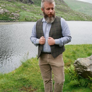 Peut inclure: Un homme portant une chemise boutonn&eacute;e bleu clair avec un gilet &agrave; carreaux brun et vert. Il se tient dans une zone herbeuse pr&egrave;s d'un lac avec une montagne en arri&egrave;re-plan.