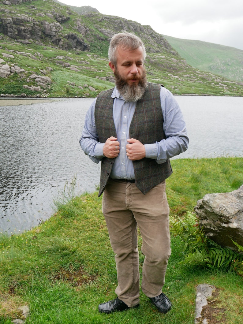 Peut inclure: Un homme portant une chemise boutonn&eacute;e bleu clair avec un gilet &agrave; carreaux brun et vert. Il se tient dans une zone herbeuse pr&egrave;s d'un lac avec une montagne en arri&egrave;re-plan.