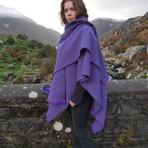 May include: A layered purple cape with a fringed edge. The cape is made of a textured fabric and is worn over a black sweater and black pants. The background features a stone wall and a mountain range.