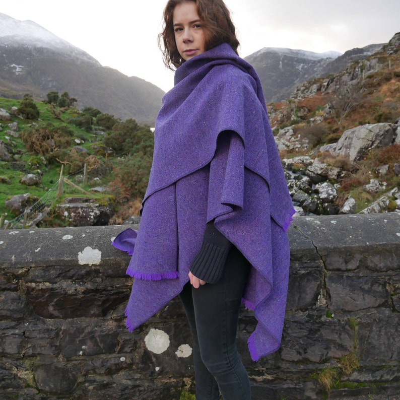 May include: A layered purple cape with a fringed edge. The cape is made of a textured fabric and is worn over a black sweater and black pants. The background features a stone wall and a mountain range.