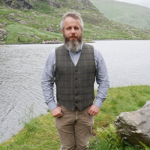 Peut inclure: Un homme barbu portant un gilet &agrave; carreaux vert et marron sur une chemise boutonn&eacute;e bleu clair. Il se tient devant un lac avec des collines verdoyantes en arri&egrave;re-plan.