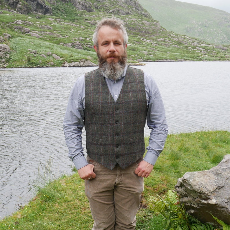 Peut inclure: Un homme barbu portant un gilet &agrave; carreaux vert et marron sur une chemise boutonn&eacute;e bleu clair. Il se tient devant un lac avec des collines verdoyantes en arri&egrave;re-plan.